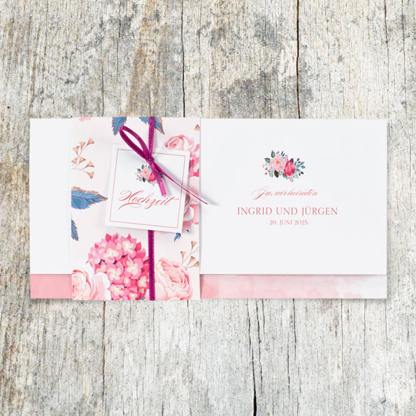 Vergrößerte Abbildung Einladungskarten zur Hochzeit, mit Banderole verschlossen, vor Hintergrund aus hellem, rustikalen Holz.